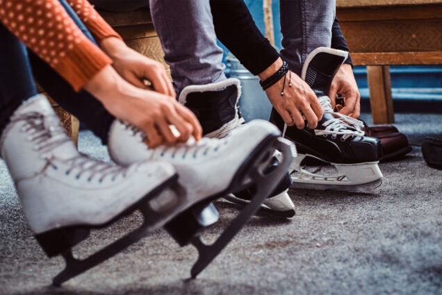 Photo: two people putting on ice skates at a hockey rink. One is lacing hockey skates and the other is lacing figure skates