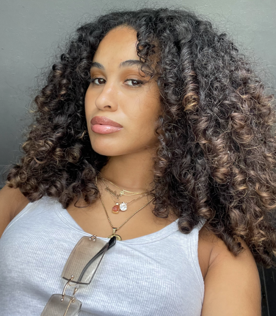 Boston University student Juliana Sena (CAS’24). This is a selfie and she has a straight face, looking at the camera, with curly hair. She is wearing a white tank top with sunglasses tucked into her top.