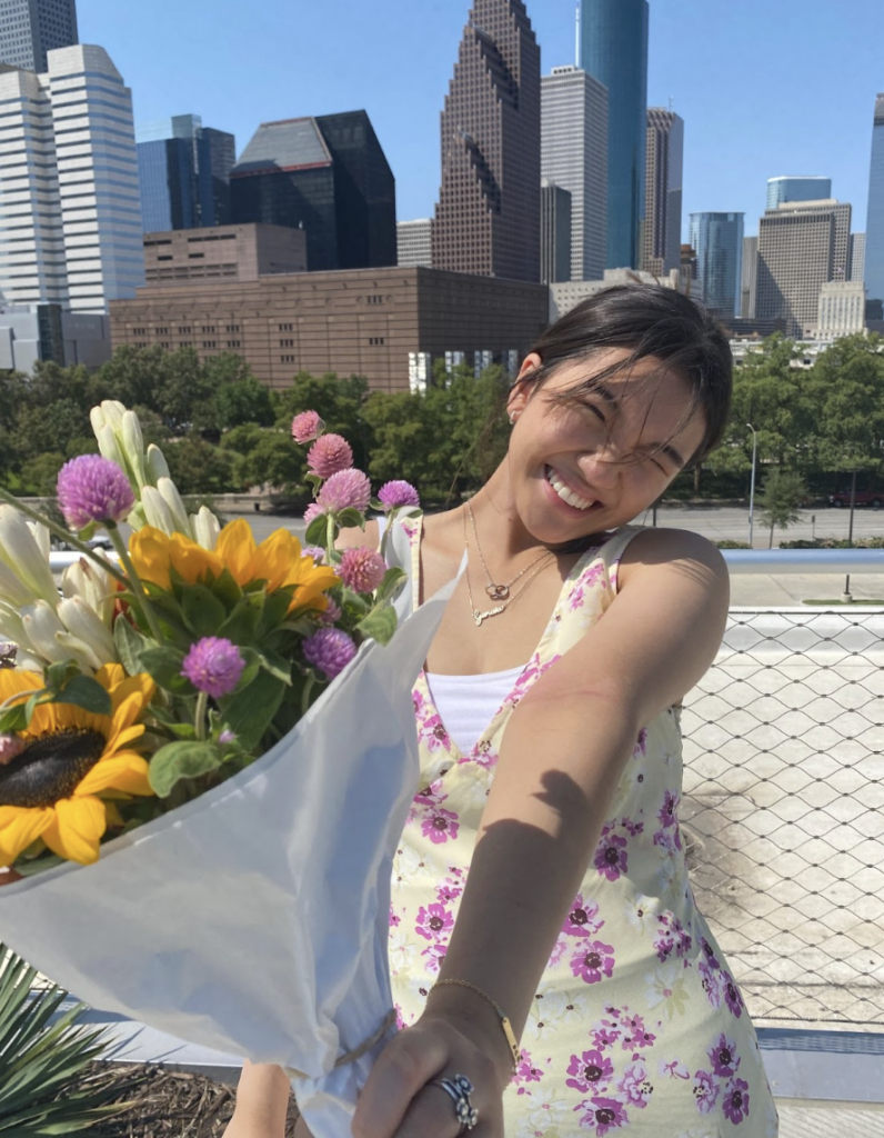 Boston University student Geneva Thai (COM ‘24). She is standing on a city side walk with tall buildings behind her. She is smiling with a floral dress on, holding a bouqet of flowers towards the camera.