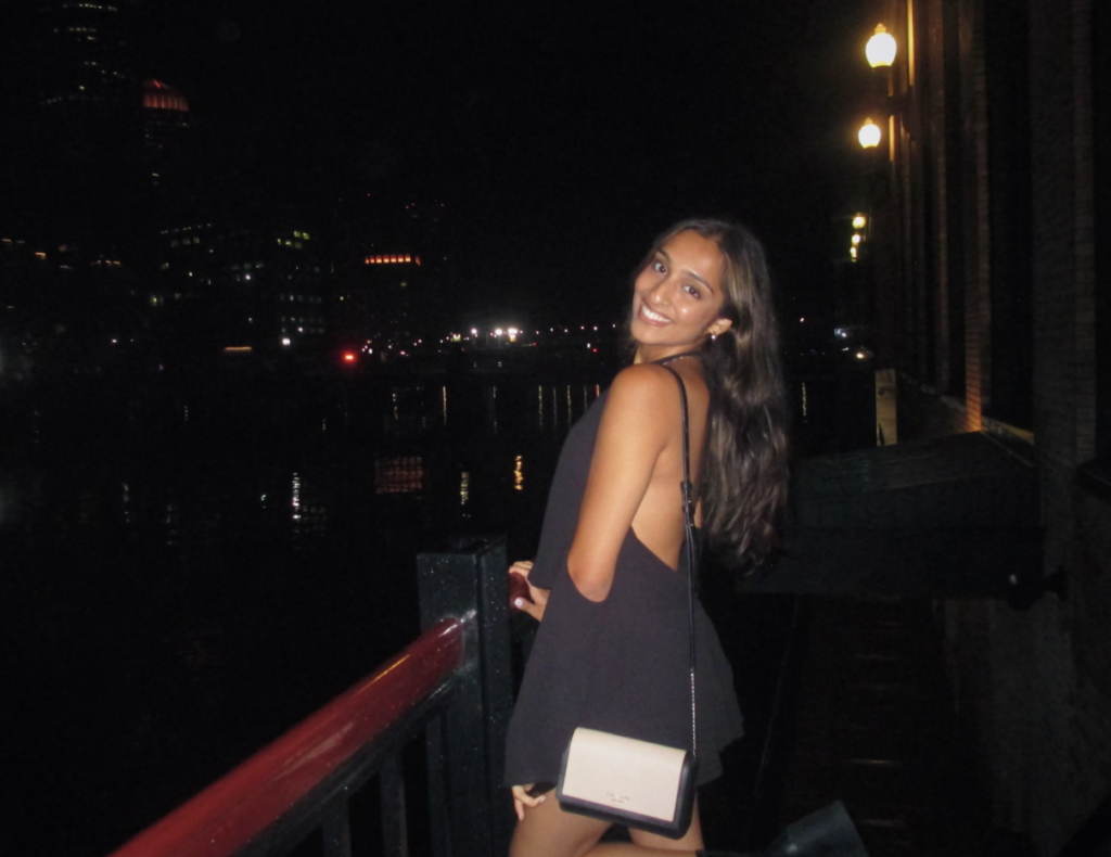 Boston University student Sajni Patel (Sargent’24). She is wearing a black dress and is outside, standing in front of a railing. The picture uses flash and she is smiling.
