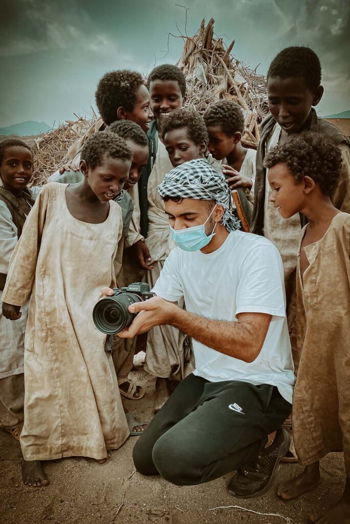 Photo: A portrait shot of a cameraman showing his photo to children from a village. The cameraman is wearing a mask and the children as excitedly looking at the camera.