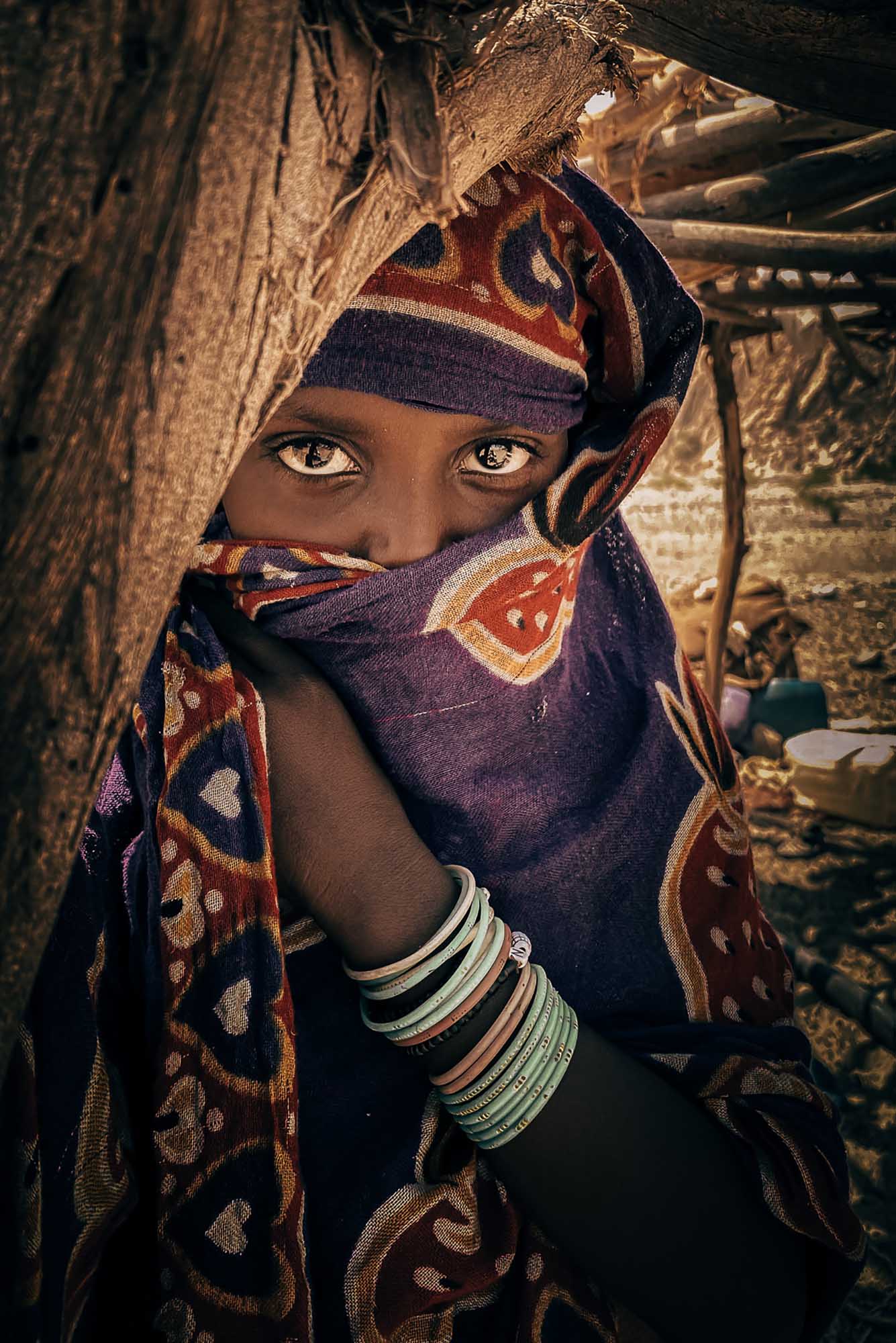 Photo: A portrait of a young individual with a scarf around their head, obstructing their view. They look through their lashes at the camera, shy and hidden around the wooden branch the lean on.