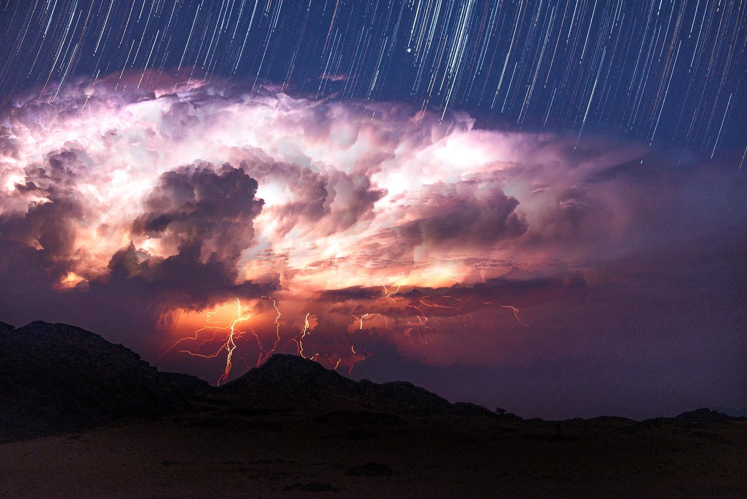 Photo: A short of a storm where, on the bottom, rain and lightening caress the land. A wall of clouds break up the viusal landscape and above the clouds, a streaky, starry sky.