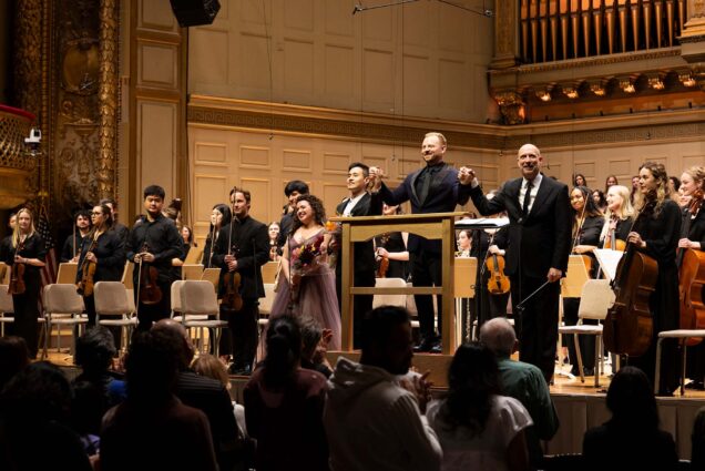 Photo: A picture of musicians on stage with some holding flowers and raising their arms as if to take a bow