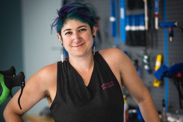 Jay Diengott, manager of BU's new Bike Kitchen, smiles for the camera. They have blue, green hair, dangling earrings, and a double nose piercing.