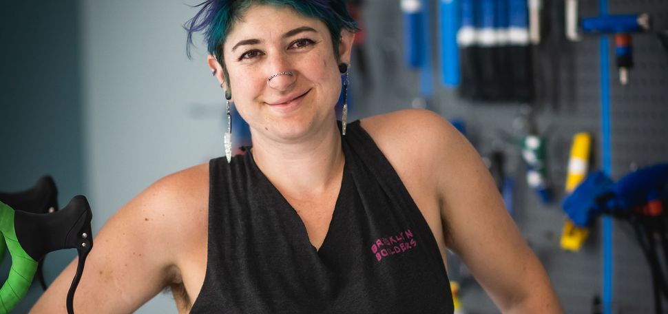 Jay Diengott, manager of BU's new Bike Kitchen, smiles for the camera. They have blue, green hair, dangling earrings, and a double nose piercing.
