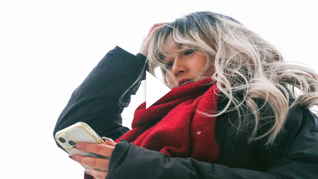 Photo: A young woman holds her head in her hand as she looks down at her phone. She has dyed blonde hair and wears a read scarf around her neck. Play button overlay.