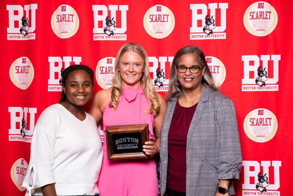 Photo: A picture of three women posing in front of a red background that says "BU." The woman in the middle is holding a plaque and wearing a pink dress