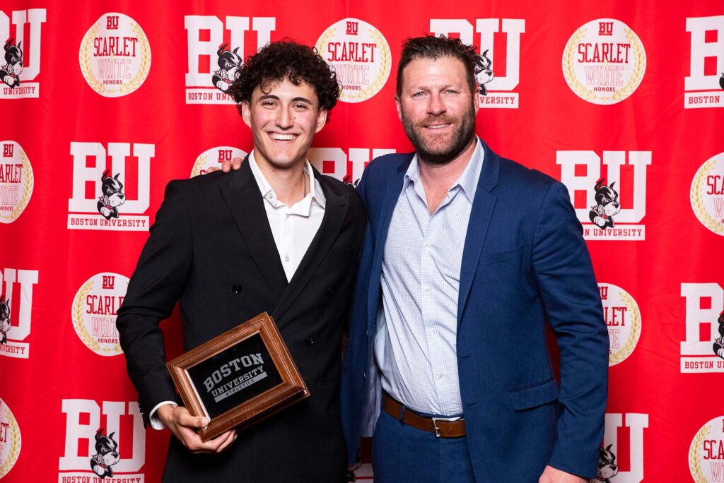 Photo: Two men in suits posing with a plaque. They are standing in front of a red background that says "BU"