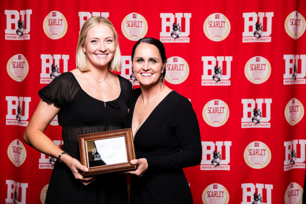 Photo: A picture of two women in black dresses posing with a plaque. They are standing in front of a red background that says "BU"
