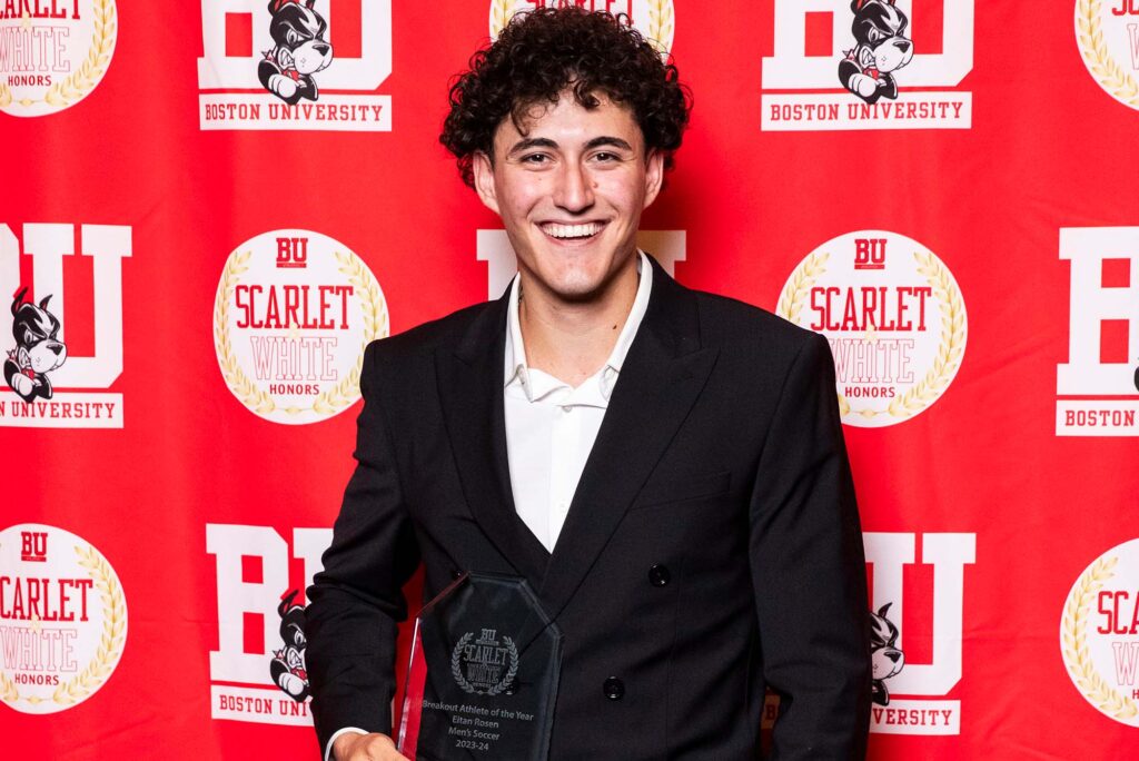 Photo: A picture of a man with curly hair wearing a black suit and posing with a clear award. There is a red background that says "BU" behind him