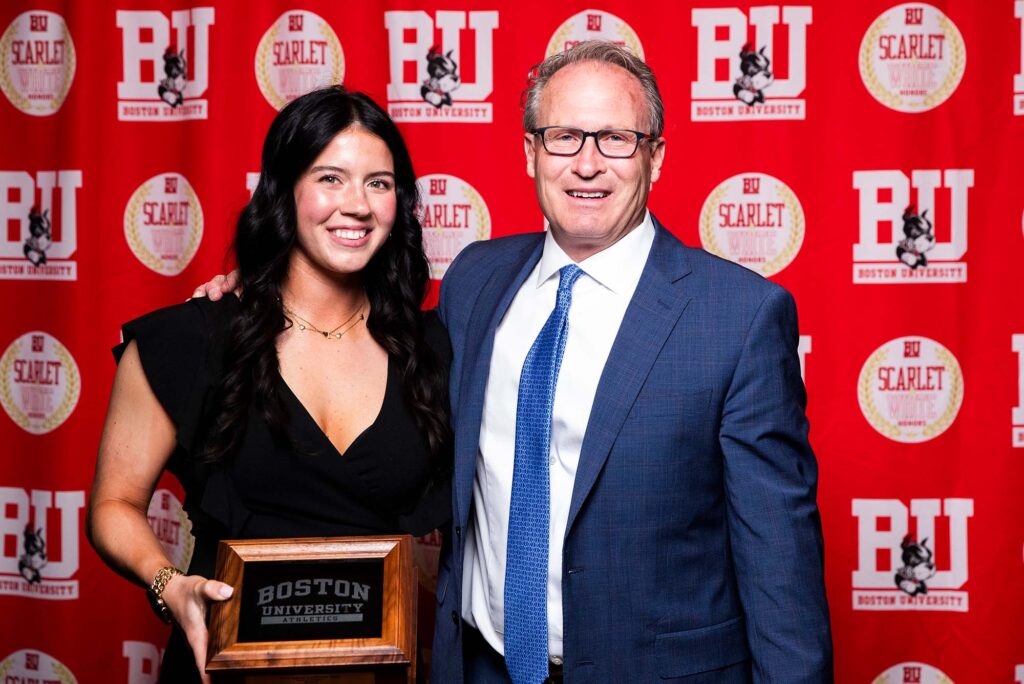 Photo: Two people posing in front of a red "BU" background. The woman on the left is wearing a black dress and holding a plaque and there is a man in a blue suit to her right