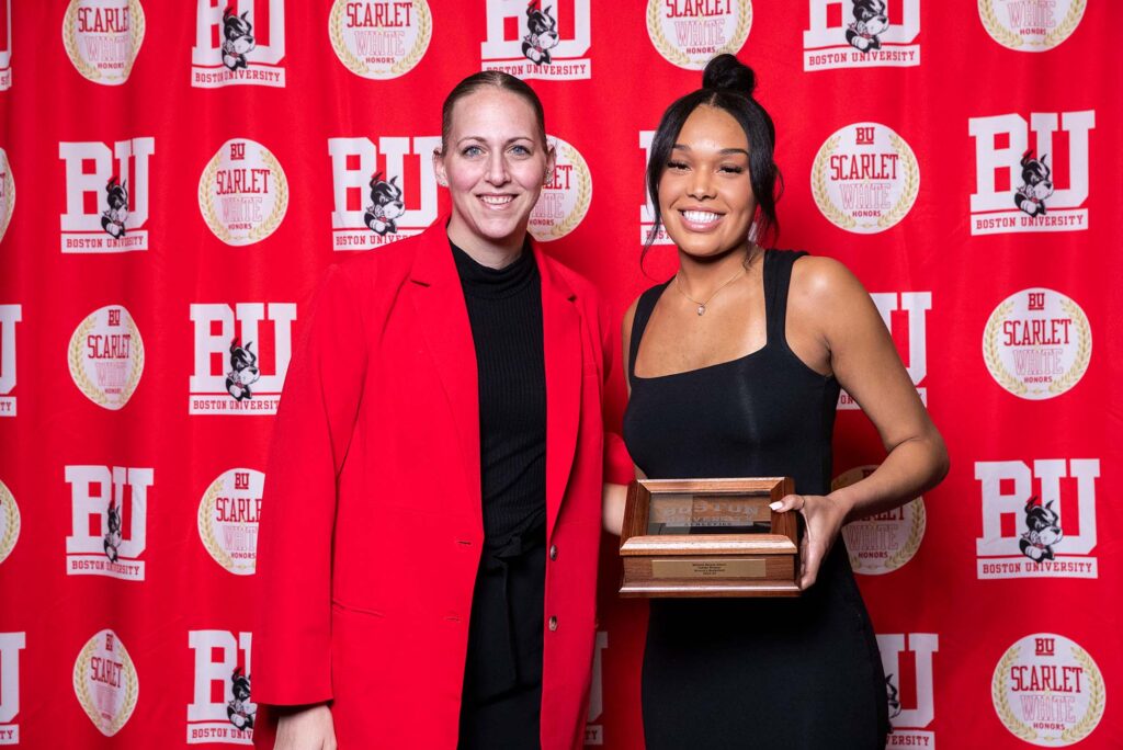 Photo: A picture of a two women posing with an award. The woman on the left is wearing a bright red jacket and the woman on the right is in a black dress. They are standing in front of a red background that says "BU"