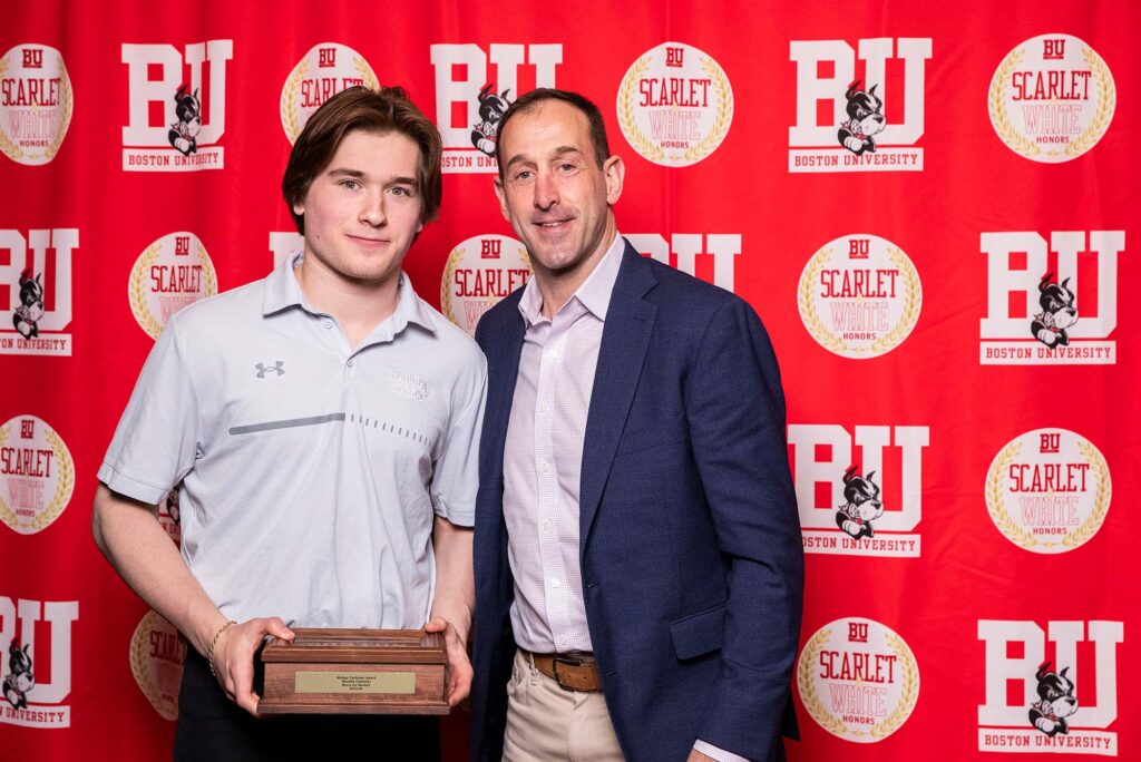 Photo: A picture of two men posing with a plaque. There is a red background that says "BU" behind them