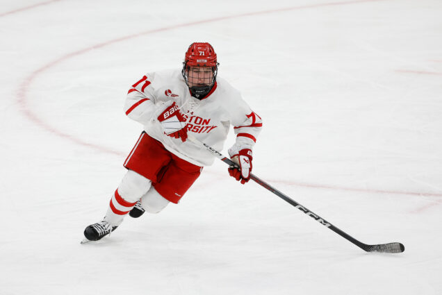 Photo: Macklin Celebrini, a BU hockey player skates on the ice in his uniform. He wears a white jersey, red shorts, and a hockey stick.