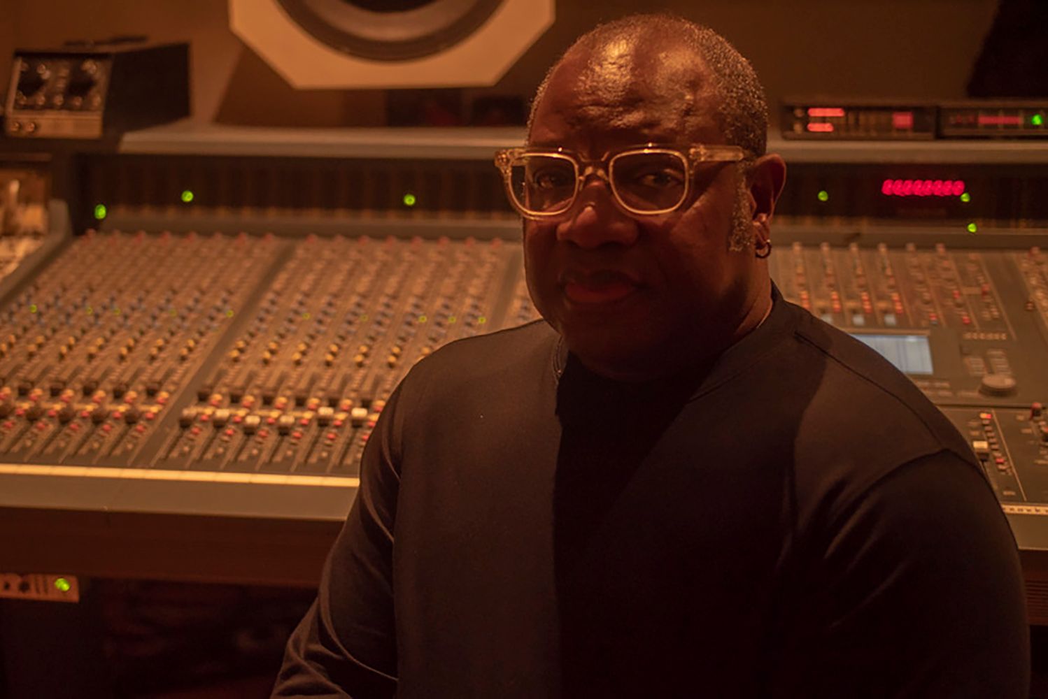 Photo: A photo with a older Black man sitting in a recording studio with the dials and controls behind him.