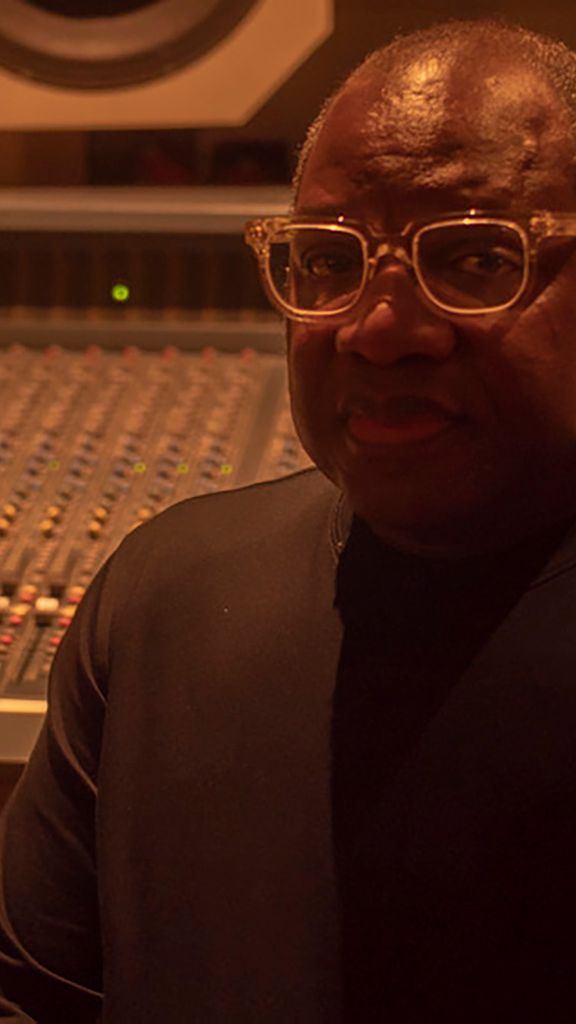 Photo: A photo with a older Black man sitting in a recording studio with the dials and controls behind him.