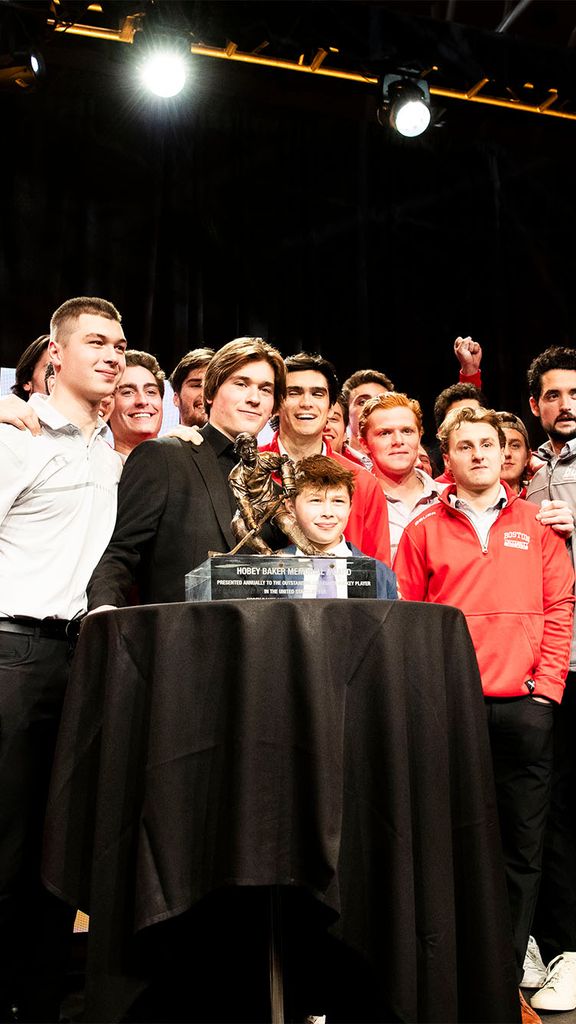 Photo: A large group of hockey players surround Macklin Celebrini who is standing behind the 2024 Hobey Baker Award trophy