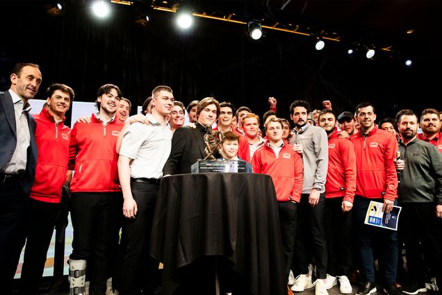 Photo: A large group of hockey players surround Macklin Celebrini who is standing behind the 2024 Hobey Baker Award trophy