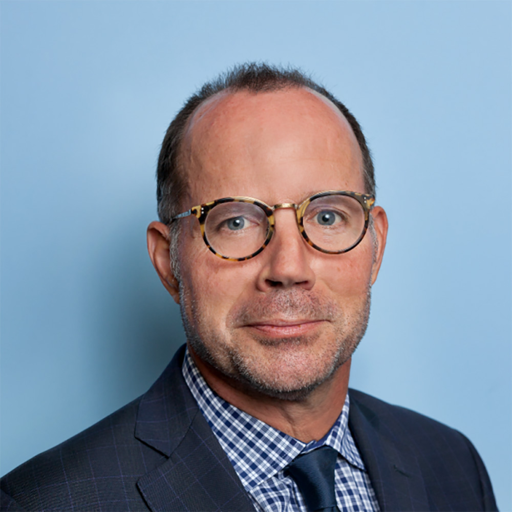 Photo: A portrait shot of a white man, Mark Williams. He poses with a suit and circular glasses in front of a blue background.