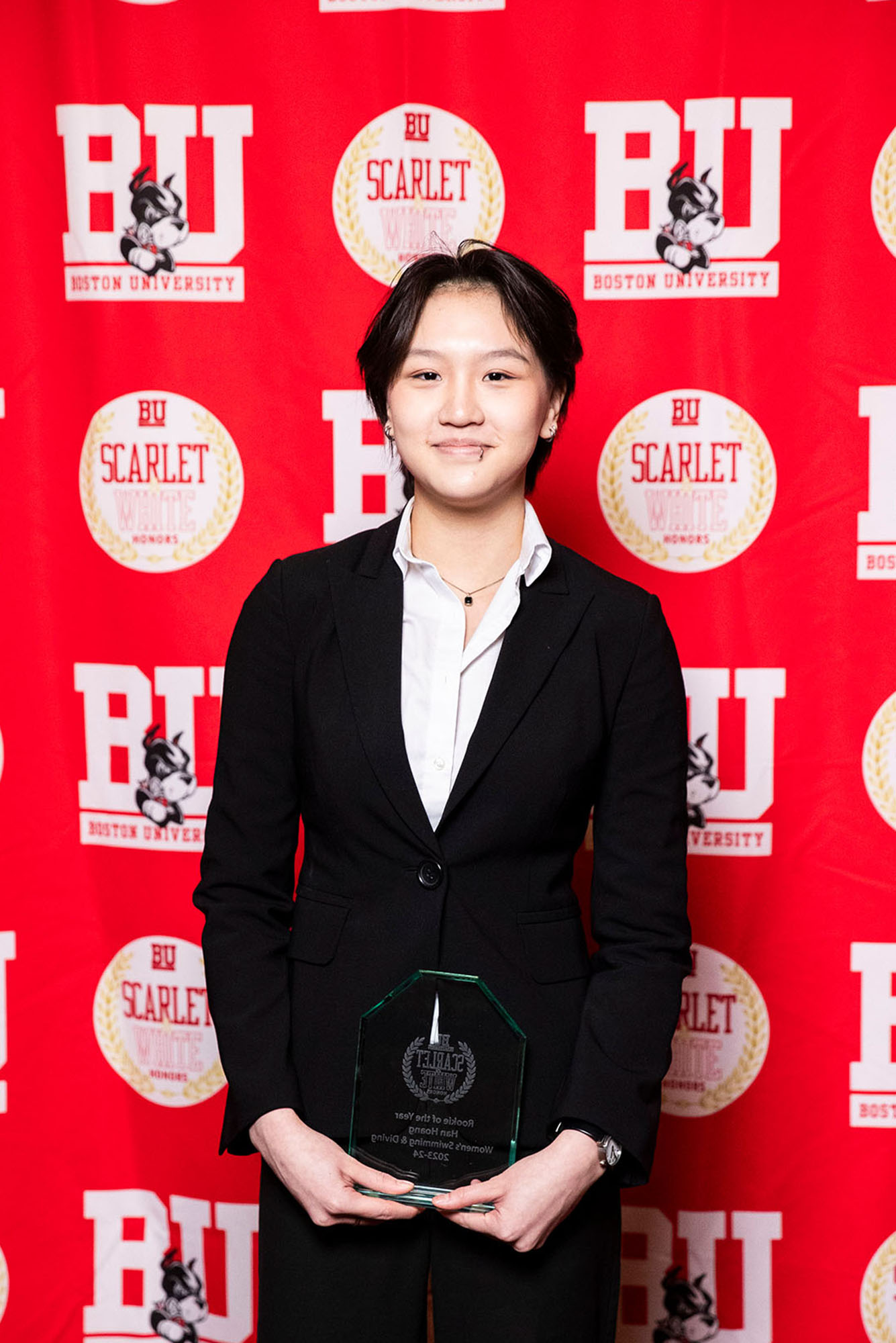 Photo: A picture of a woman in a black suit holding a clear award. There is a red background that says "BU" behind her