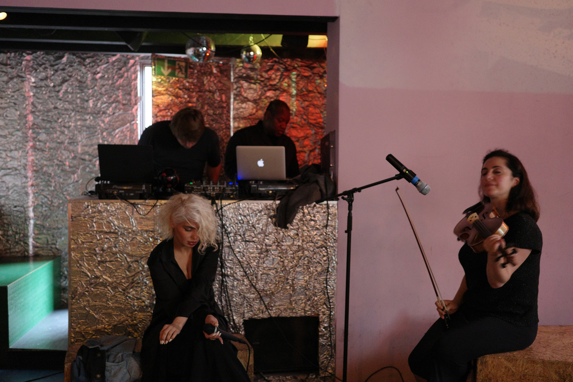 Photo: A small studio scene with a individual sitting by a mic on the right hand side. In the center, another individual with a funky haircut sits in front of another microphone.