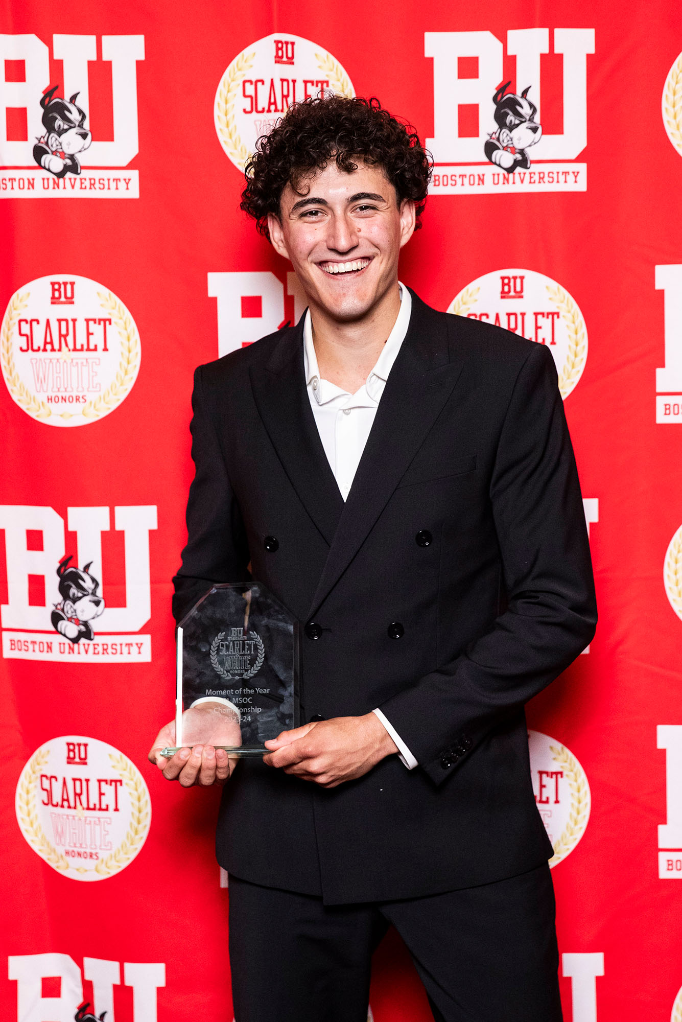 Photo: A picture of a man with curly hair wearing a suit and holding an award. There is a red background behind him that says "BU"