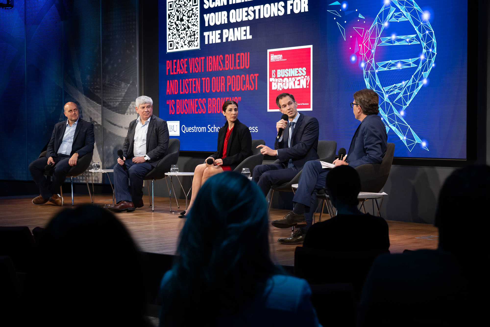 Photo: A panel of five people sitting in a row on a stage in front of a crowd wearing formal attire