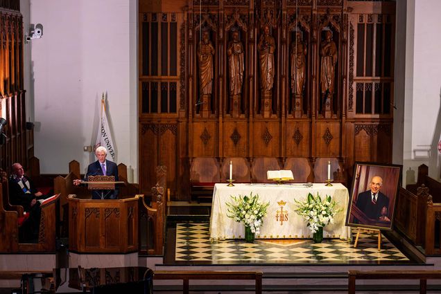 Photo: A shot of Aram Chobanian's memorial service. A picture of Aram Chobanian rests on the right hand side while a speaker stands to the left. A table with cloth and flowers sits in between them.