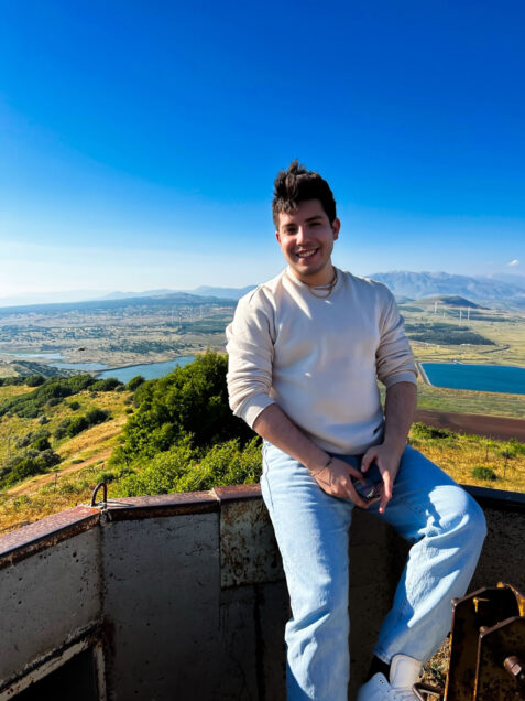 Photo: Michael Arellano, a young individual with short black hair wearing a white crew neck, sits casually on a stone wall overlooking a vast landscape. They smile for the photo.