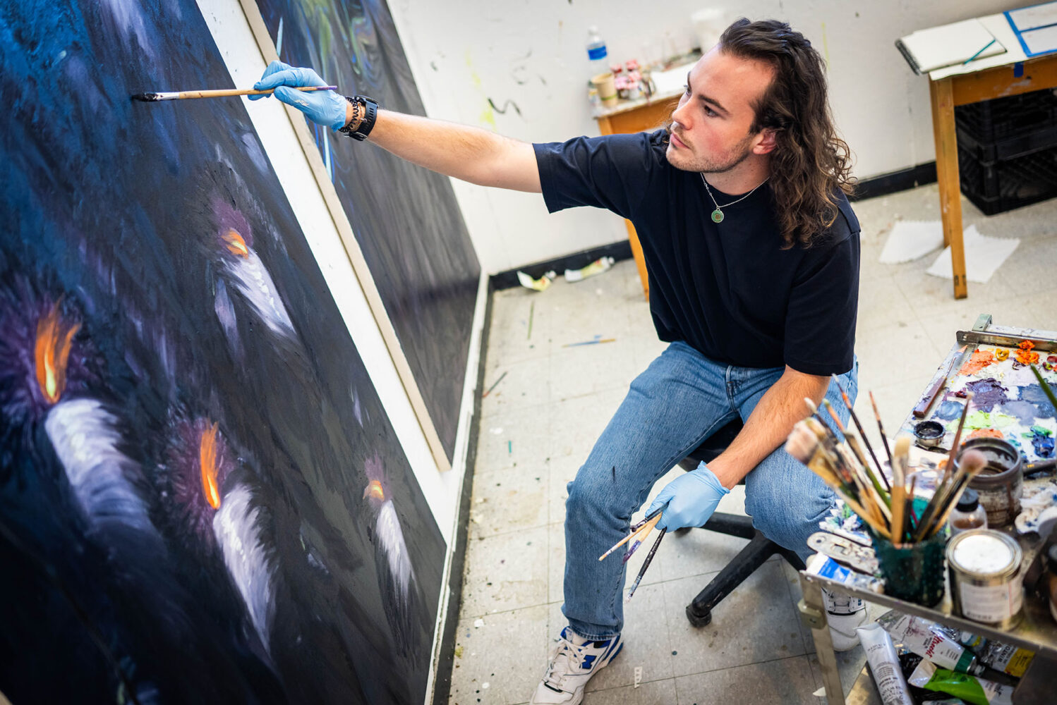 Photo: A young man with long hair wearing a black tshirt and jeans, lifts his brush up to paint on a canvas.