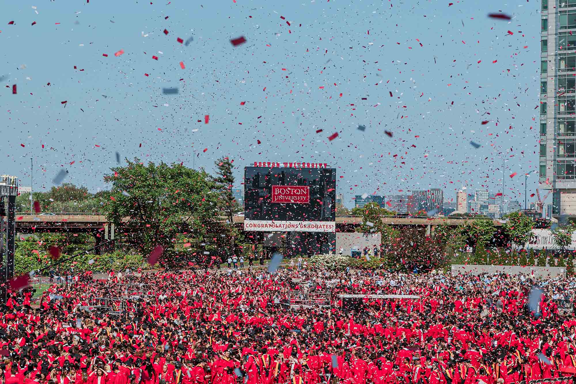 Photo: Boston University’s 150th Commencement. Confetti flies and falls through the air as graduates celebrate their accomplishments. A big screen with BOSTON UNIVERSITY on the front stands in the middle.