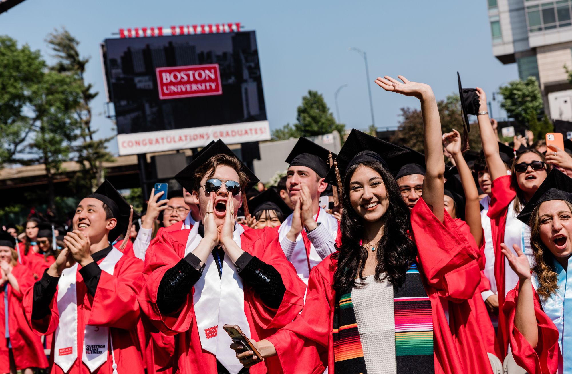 A celebratory scene at Boston University with a group of graduates in red robes and black caps, joyfully raising their hands and capturing photos under a clear blue sky. They are standing in front of a large billboard that reads ‘BOSTON UNIVERSITY CONGRATULATIONS GRADUATES!’ in bold white letters on a red background, marking the festive occasion of their graduation.