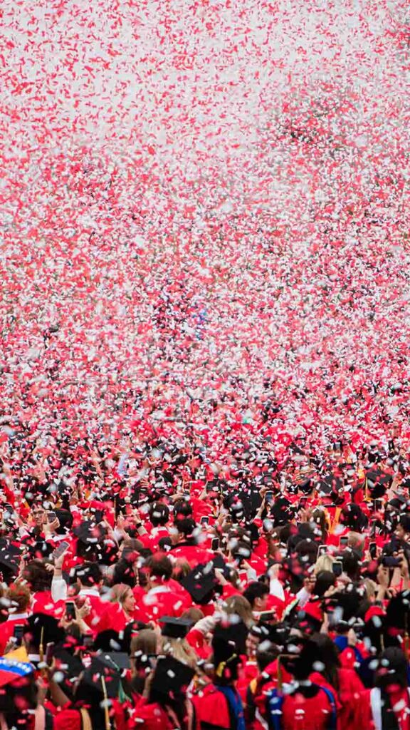 Photo: a field full of Boston University graduates wearing red gowns and black caps, with confetti falling all around