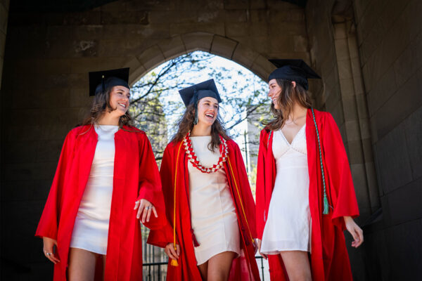 Photo: Three college graduates in caps, red gowns, and white dresses smile at each other as they walk toward the camera