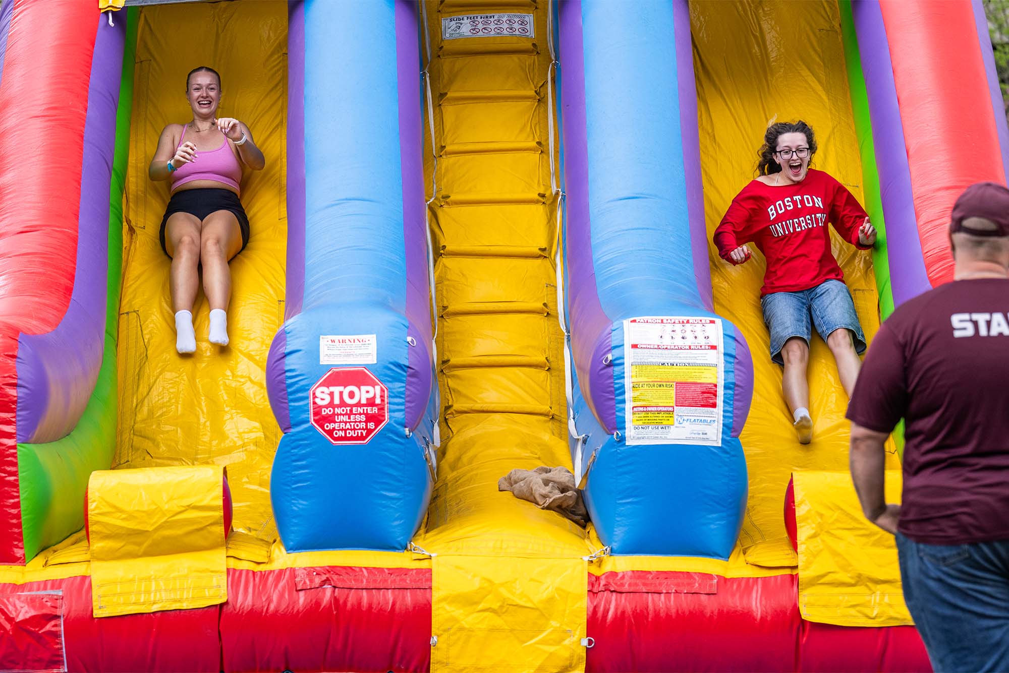 Photo: Two college students slide down a large inflatable slide. Jessica Spiers (CAS’24), in pink, and Reghan Lanning (SAR’24) launch down the inflatable slide at Senior Carnival on BU Beach May 15. The event, which is part of Senior Week, offered cotton candy, bbq, lawn games, drinks, and inflatables. Photo by Cydney Scott