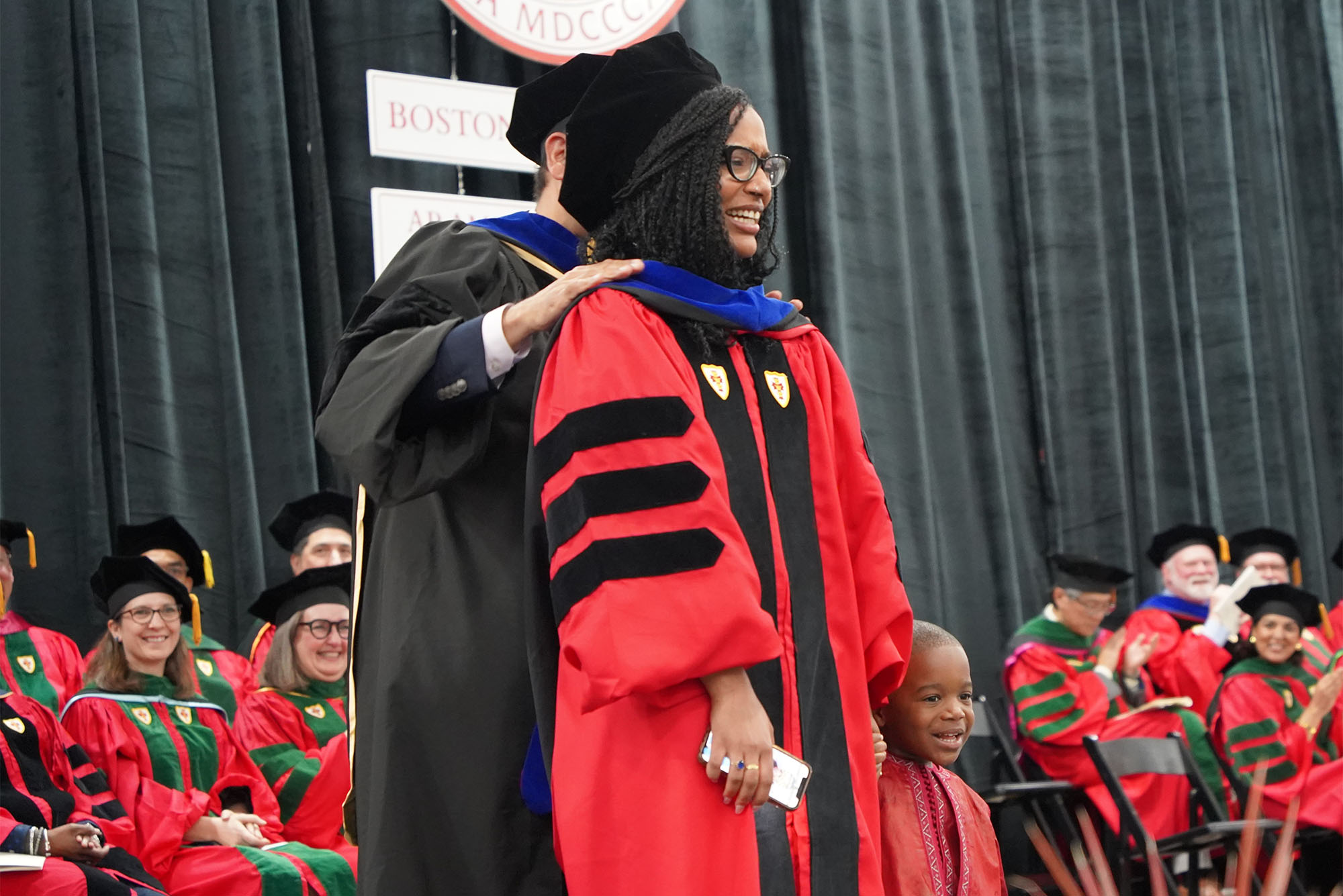 Photo: MD student speaker Josiane Fofana (CAMED’24), with her three-year-old, as she receives her doctoral hood at the BU medical school’s MD and PhD convocation ceremony May 16. A black woman wearing a robe and doctoral hood stands in front of a room of other graduates who are clapping