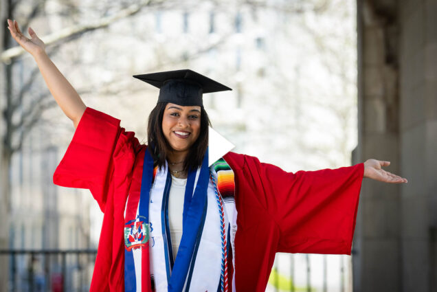 Photo: A college student in a red robe with black mortarboard and blue tassles holds their arms up in celebration. A white play button overlaid on top of the image