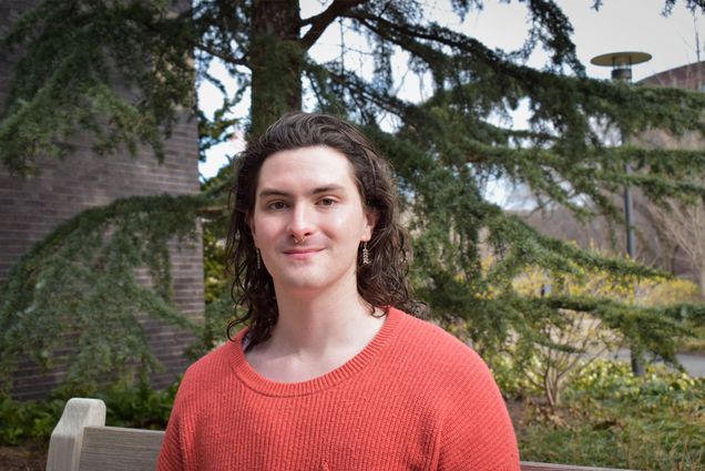 Photo: A person with short dark hair and a nose ring wearing an orange shirt in front of a verdant nature scene with pine trees