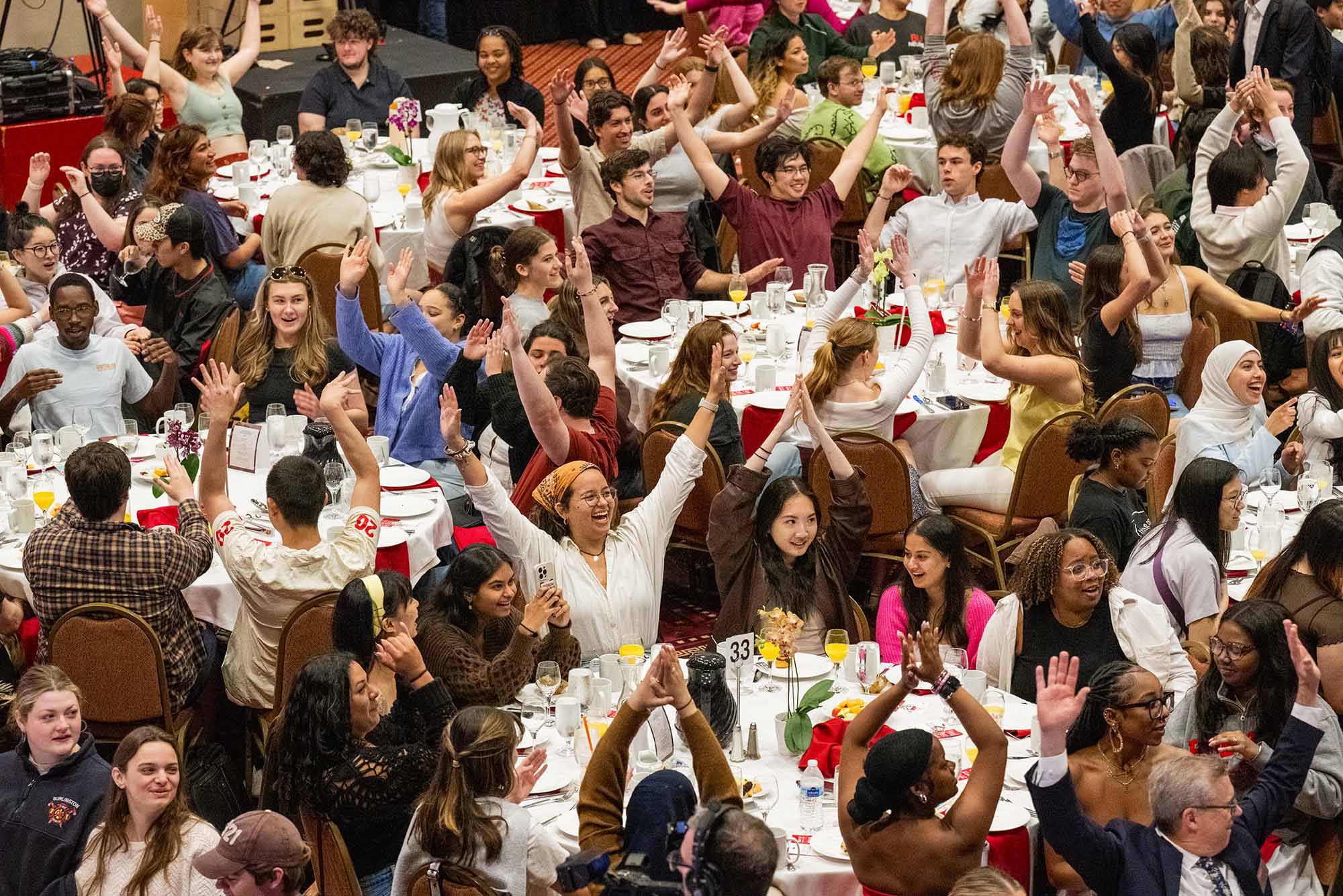 Photo: A picture of many people celebrating with their arms raised as they sit around round tables