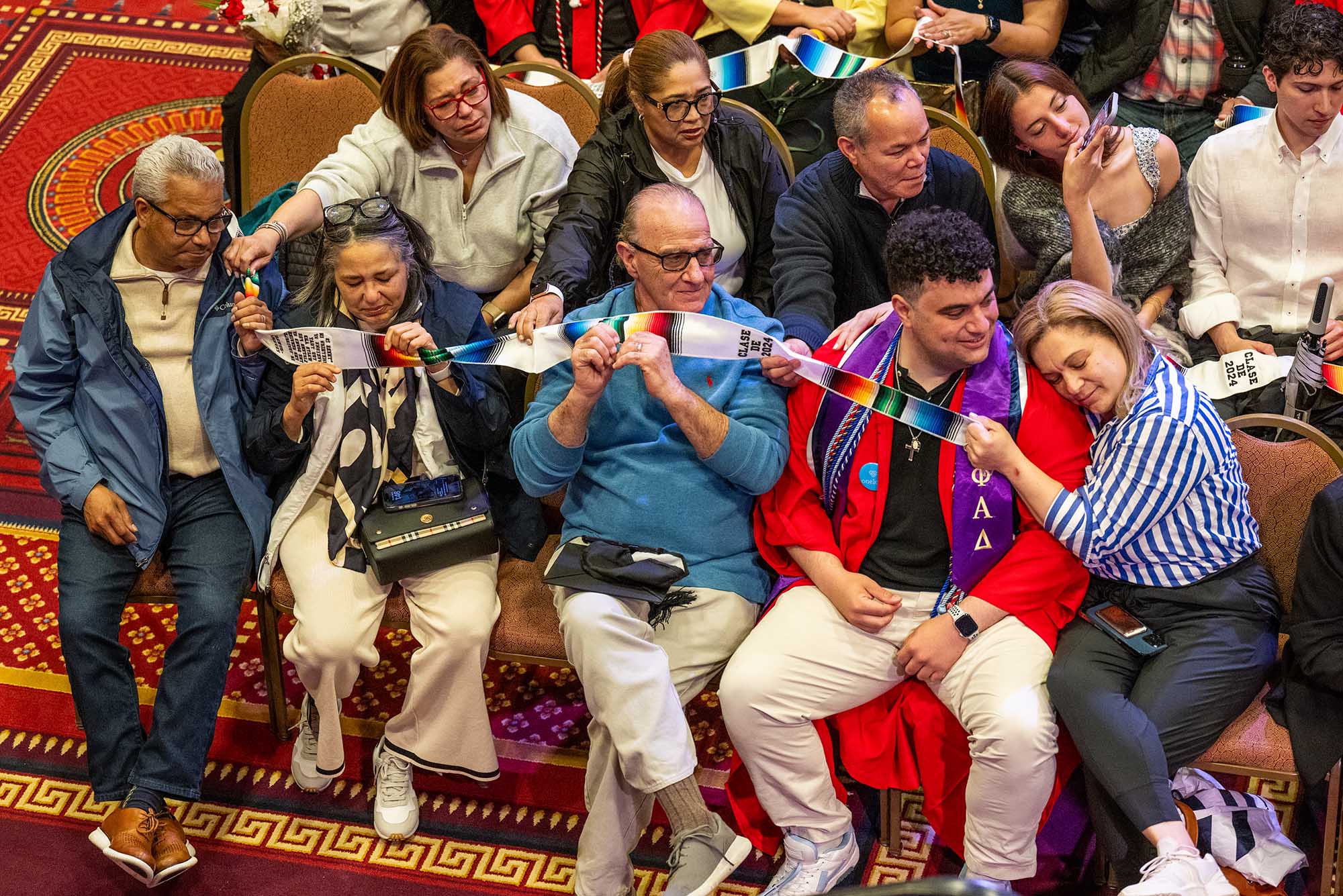 Photo: A large family sits in rows of chairs as they hold a banner indicating a first-gen senior at BU. Their faces are sentimental, filled with pride and joy, and turned to look at Andrew Cili, the student getting the honor. His mom, on his right, cuddles into his arm in admiration and love.