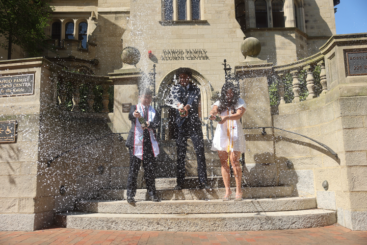 Three individuals in graduation wear standing on the steps of Boston University's alumni center, spraying champagne bottles in front of themselves. 
