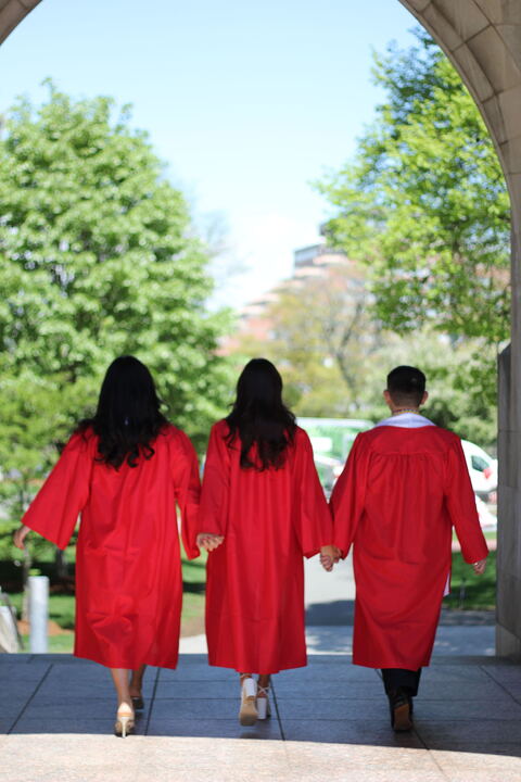 Three Boston University students in red graduation gowns holding hands and facing their backs toward the camera. In front of them is a green field with trees in full summer bloom.