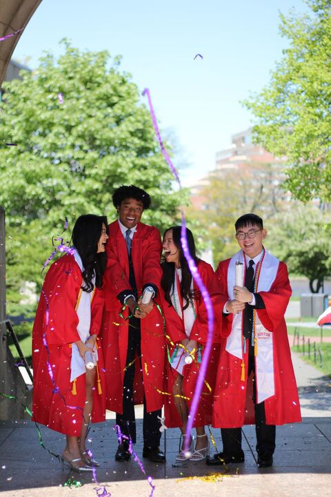 Boston University students celebrating their graduation. They are in red gowns and spraying confetti at the camera. 