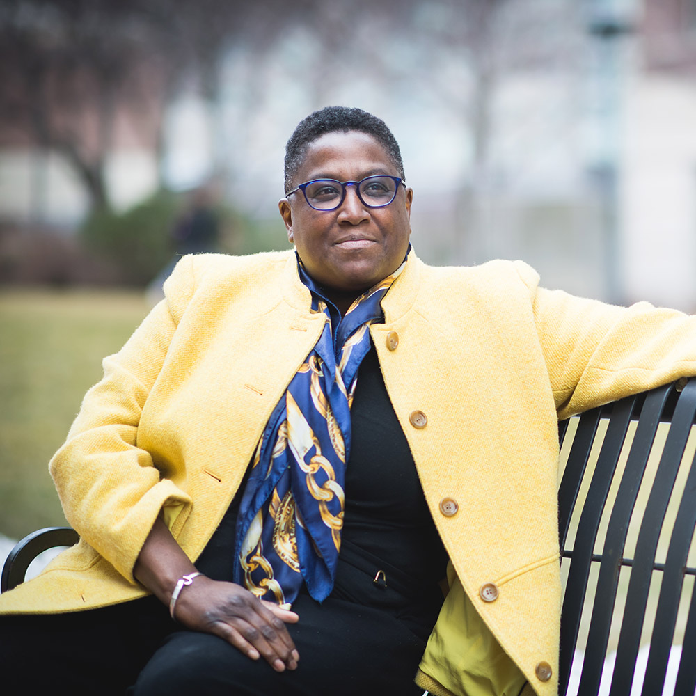 Photo: Judi Burgess, a Black woman with a black shirt, blue and gold scarf, and yellow jacket blazer, pose for a photo. She is the director of Labor Relations for BU and co-director of BU's LGBTQIA task force.