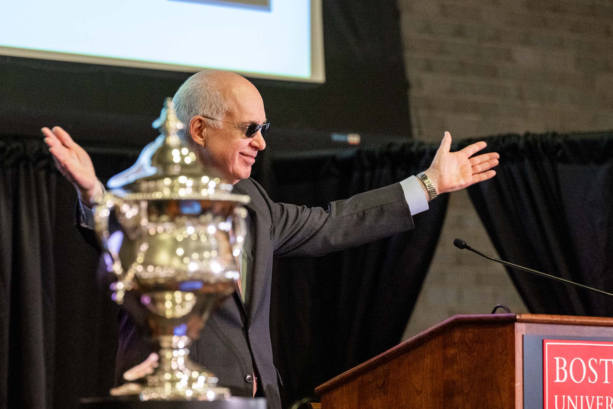 Photo: A picture of a man with white hair in sunglasses raising his arms behind a podium