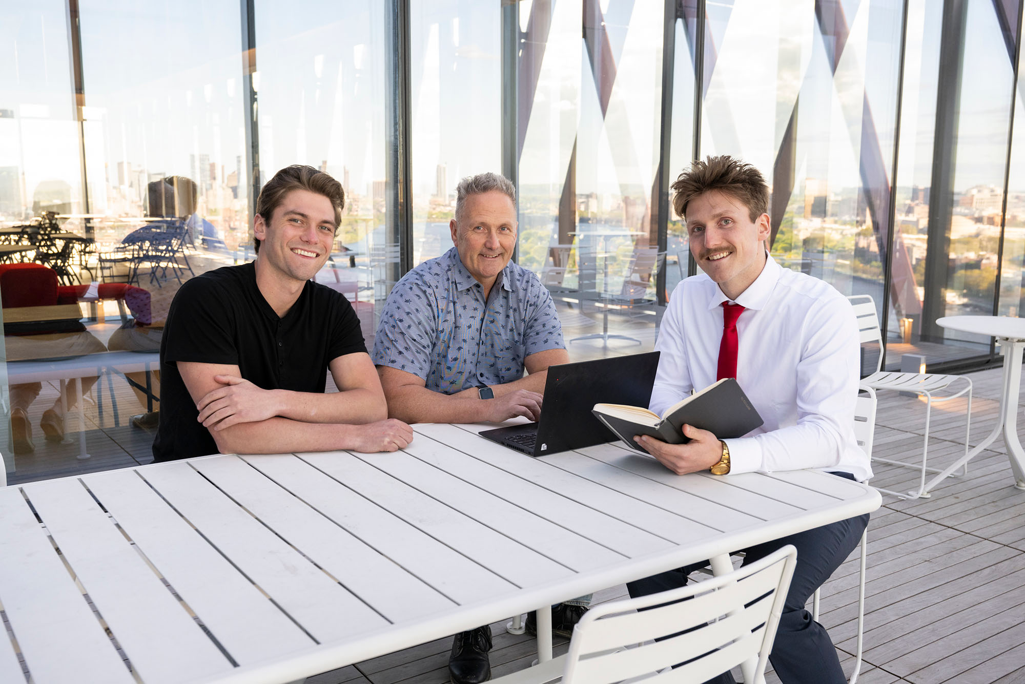 Photo: A picture of a father sitting at a white outdoor table with his sons on either side of him. The father has a laptop open in front of him and the son to his right has a book open