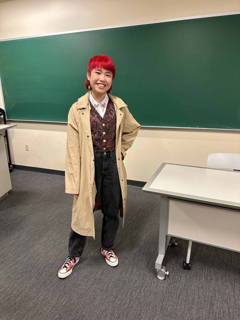 Photo: A picture of a person with short, bright red hair and a long tan coat. They are also wearing a patterned vest with buttons that is tucked into black jeans
