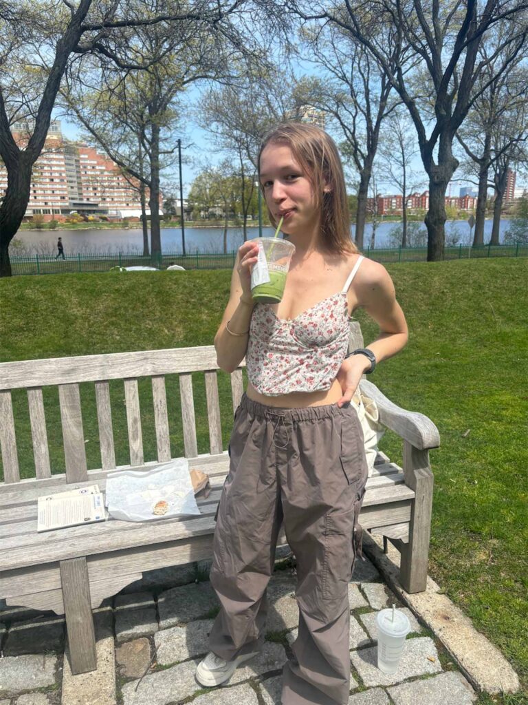 Photo: A picture of a girl with light, short hair sipping a matcha as she poses for a photo. She is wearing dark tan cargo pants and a floral tank crop top