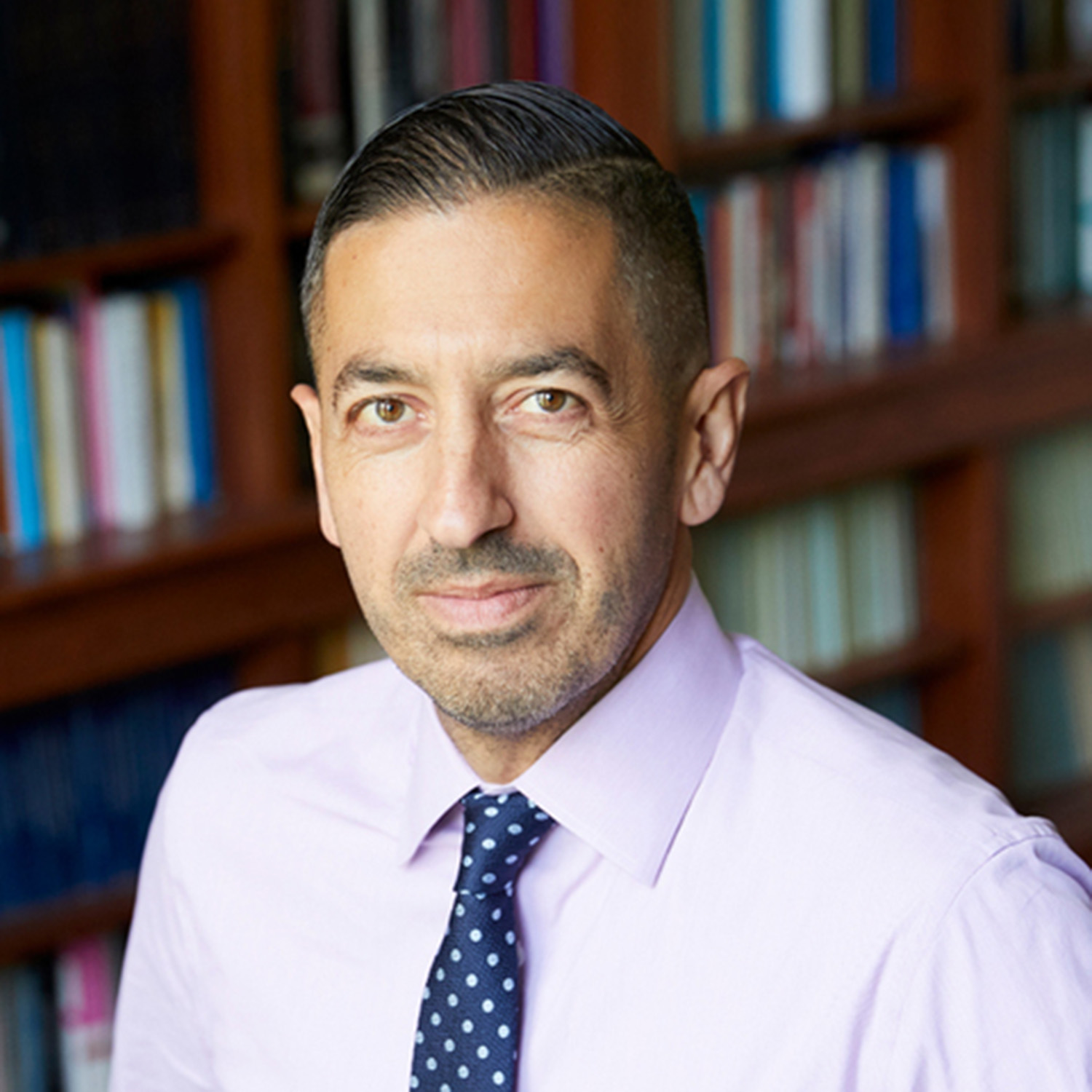 Photo: A headshot of a man with dark hair wearing a light pink shirt and blue tie with white polka dots. He in posing in front of a bookshelf